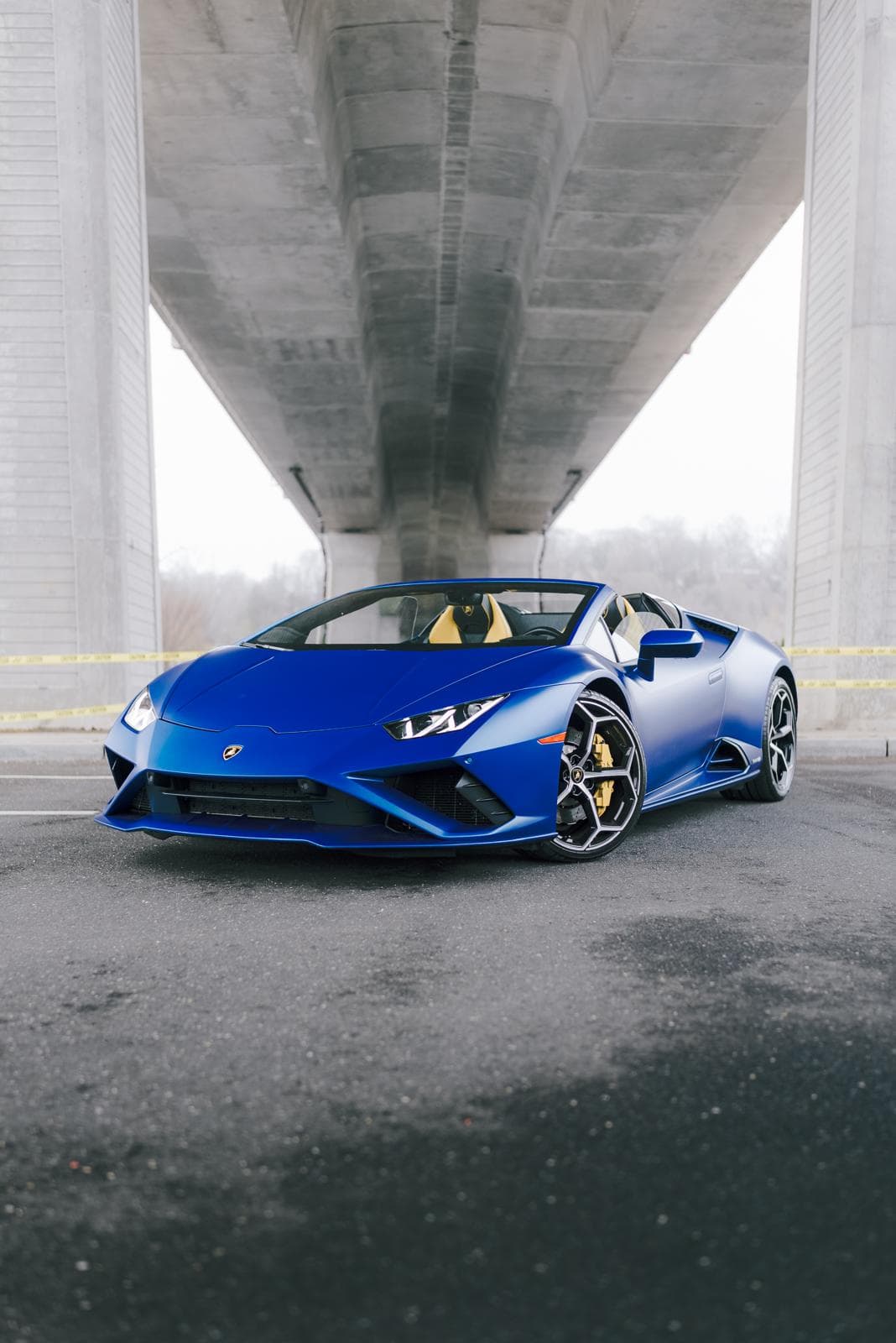 Matte blue Lamborghini Huracán EVO Spyder seen head-on between the pillars of a bridge, yellow seats visible