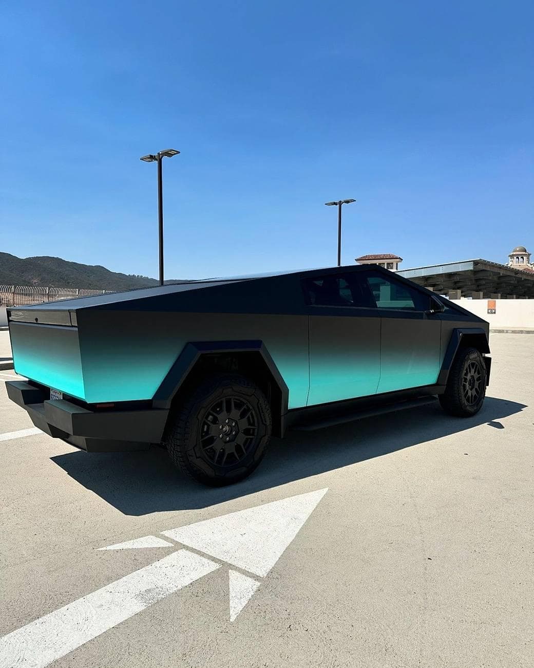 Tesla Cybertruck wrapped in a black-to-teal fade, rear three-quarter view on a rooftop parking deck